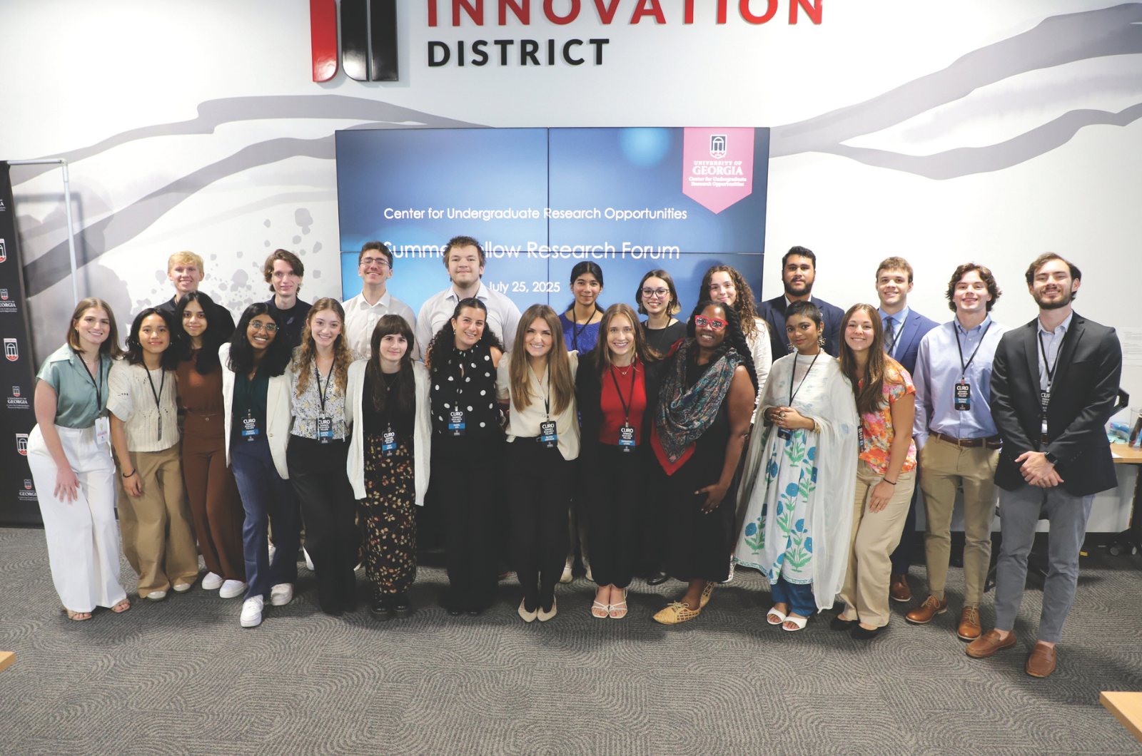 A group of about 20 CURO Summer Research Fellows and staff pose together in front of a screen reading "Center for Undergraduate Research Opportunities: Summer Fellow Research Forum, July 25, 2025" inside UGA's Innovation District.