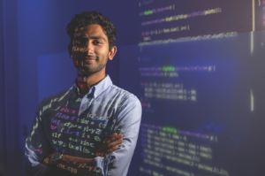 Sudhan Chitgopkar stands with arms crossed as lines of green and pink computer code are projected across his face and shirt, with a screen of code visible in the background.