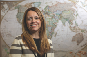 Maryann Gallagher smiles in front of a large world map, wearing a striped cream and gray cardigan.
