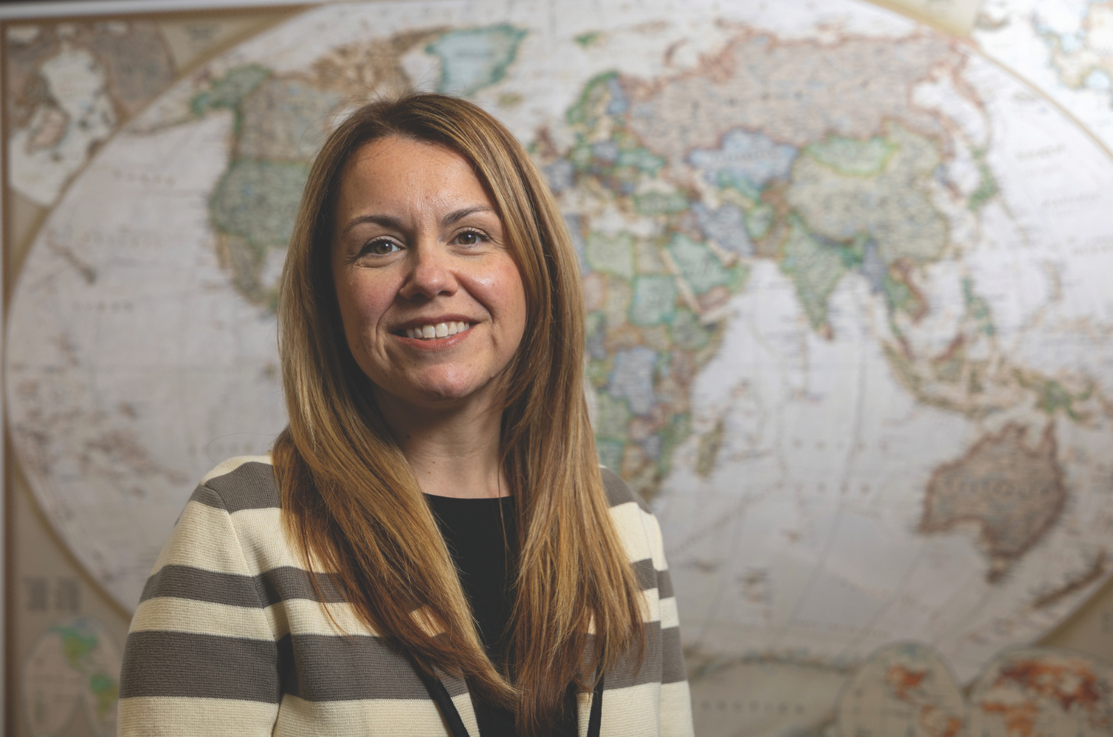 Maryann Gallagher smiles in front of a large world map, wearing a striped cream and gray cardigan.